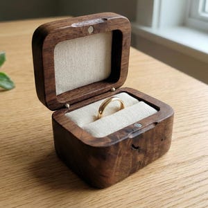 May include: Open wooden ring box with a gold ring inside. The box is square with rounded edges and a magnetic closure. The interior is lined with a light-colored fabric. The box is sitting on a wooden surface.