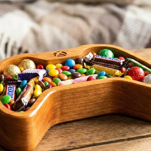 May include: A wooden candy dish shaped like a video game controller, filled with colorful candies and wrapped chocolates. The dish is made of light-colored wood and sits on a wooden table. The candies include M&Ms, Snickers, and other treats.