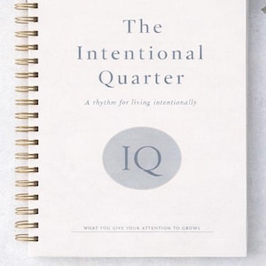 Pode incluir: Um caderno espiralado creme com o título "The Intentional Quarter" em letras cinzentas. A capa também inclui o texto "A rhythm for living intentionally" e "WHAT YOU GIVE YOUR ATTENTION TO GROWS". Um círculo cinzento com as letras "IQ" também está na capa.