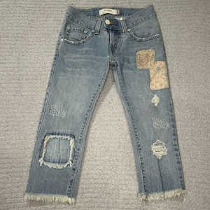 May include: Light blue denim jeans with distressed details, including patches and frayed edges. The jeans feature decorative patches on the right leg and a raw hem. The jeans have a button and zipper closure.