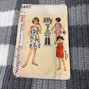 Può includere: Una busta per cartamodello vintage Simplicity, numero 5467, per un abito Jiffy in taglia 12. La parte anteriore presenta illustrazioni di tre diversi stili di abito, tra cui una stampa floreale, righe verticali e un design arancione a tinta unita.