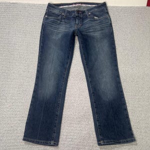 Puede incluir: Un par de pantalones vaqueros de mezclilla azul oscuro con un diseño clásico de cinco bolsillos, cierre de botón y cremallera, trabillas para cinturón y un desvanecido sutil en los muslos y rodillas, con corte recto y costuras visibles.