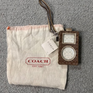 May include: A brown and tan monogram print ID holder with a wrist strap and a white dust bag featuring the red Coach logo and text EST. 1941. A white Coach Factory tag is attached to the strap, and the holder has a rectangular and circular window.