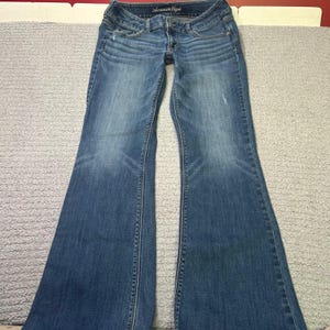 May include: Blue denim flared jeans with a faded wash. The jeans have a classic five-pocket design, a zip fly, and a button closure. The brand name "American Eagle" is visible on the waistband.