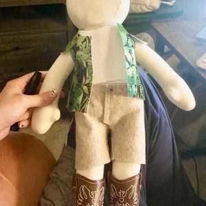 以下が含まれることがあります： 白いシャツ、ベージュのショートパンツ、緑の模様のベストを着た、顔が空白の手作りの人形。人形は、白いステッチと茶色の巻き毛が付いた茶色のカウボーイブーツを履いています。
