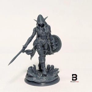 May include: A grey unpainted resin miniature of a hooded elf warrior standing on a textured base. The figure wears armor, a cloak, and holds a sword in the right hand and a round shield in the left. A small logo with the letter B is visible.
