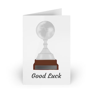 May include: A white greeting card featuring a silver trophy design. The trophy has a globe at the top, a silver stem, and a brown base. The words "Good Luck" are written in black script at the bottom.