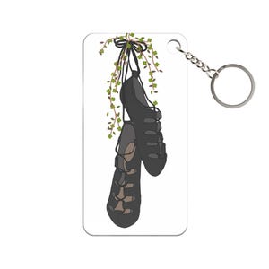 Puede incluir: Un llavero rectangular con un gráfico de dos zapatos de baile irlandés negros colgando de una vid verde con tréboles. El llavero tiene una cadena y un anillo plateados.