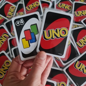 Może przedstawiać: Dwie karty Uno są trzymane przed stosem innych kart Uno. Jedna karta ma czarne tło z czerwonym logo "UNO". Druga karta ma białe tło z symbolem "+4" i kolorowymi kwadratami.