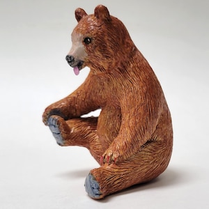 Op de afbeelding: Een bruine berenfiguur met gestructureerde vacht, zittend in een achteroverleunende positie met één poot omhoog. De beer heeft een lichtgekleurde snuit, een klein roze tongetje dat uit de bek steekt, grijze voetzolen en rood geschilderde klauwen.