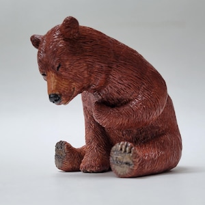 Op de afbeelding: Een klein bruin berenbeeldje in zittende houding met het hoofd naar beneden gebogen. De beer heeft gedetailleerde vachttexturen, een zwarte neus en gesloten ogen, met een voorpoot rustend tegen de borst in een peinzende houding.