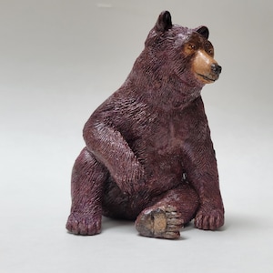 May include: A dark brown bear figurine in a seated position with a textured fur finish. The bear has a lighter tan-coloured snout, a black nose, and dark eyes, with its head turned slightly to the right. The sculpture shows detailed fur.