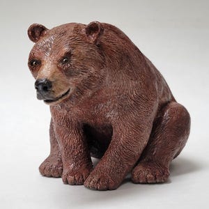 May include: A brown bear figurine with a textured fur finish, depicted in a seated position. The bear has a dark nose, small rounded ears, and a focused expression, with detailed sculpting that mimics the appearance of thick, coarse animal fur.