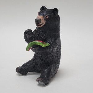 Könnte beinhalten: Eine schwarze Bärenfigur in sitzender Position mit strukturierter Fellzeichnung und einer hellbraunen Schnauze. Der Bär hält einen gebogenen, grün-rosa Fisch in seinen Vorderpfoten, wobei der Kopf leicht nach links gedreht ist.