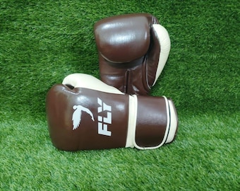 May include: A pair of brown and cream-colored boxing gloves. The gloves have a white bird logo and the word "FLY" printed on the side. The gloves are set against a green background.