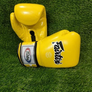 Könnte beinhalten: Gelbe Boxhandschuhe mit dem schwarzen Schriftzug "Fairtex" auf der Seite. Die Handschuhe bestehen aus glänzendem Material. Der Hintergrund ist grün.