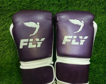 May include: A pair of purple boxing gloves with white accents. The gloves feature the word "FLY" in white block letters and a white wing logo. The gloves are set against a green background.