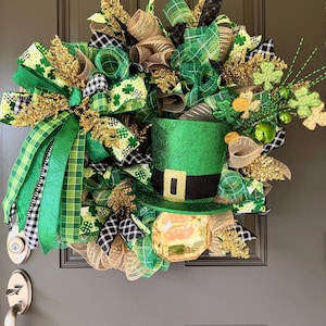 May include: A St. Patrick's Day wreath featuring a green top hat with a black band and gold buckle. The wreath is adorned with green, gold, and black ribbons, shamrocks, and decorative accents, creating a festive holiday decoration.