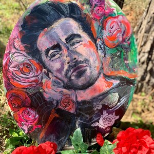 May include: A round painting featuring a portrait of a man surrounded by roses. The artwork uses a palette of pinks, reds, and blacks. The man's face is painted with a thoughtful expression. Red roses and green foliage are in the foreground.