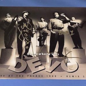 Puede incluir: Portada de álbum en blanco y negro con la banda Devo, con firmas. Los miembros de la banda están de pie sobre pedestales. El título del álbum es "Somewhere with Devo" y "Live at the Palace 1988 + Remix Set" está impreso en la parte inferior.