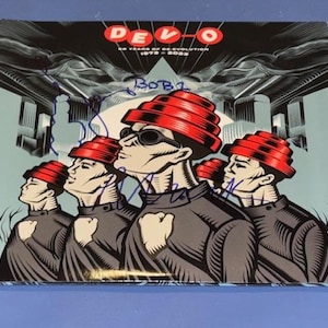 Può includere: Copertina dell'album con un'illustrazione stilizzata della band Devo. L'immagine mostra cinque figure con cappelli a cupola energetica rossi e camicie nere. La copertina dell'album presenta il testo "DEVO" e "40 Years of Re-volution 1973-2023".