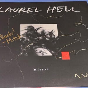 May include: Black album cover with the text "LAUREL HELL" in white script. A black and white photo of a person's face and hair is in the center. The name "mitski" is at the bottom. Gold script says "thanks! -Mitski".