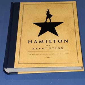 May include: A hardcover book titled "Hamilton: The Revolution" by Lin-Manuel Miranda and Jeremy McCarter. The cover features a black silhouette of a person on a star, with the title in gold lettering on a tan background.
