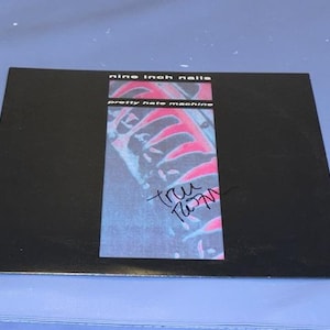 May include: A black album cover for Nine Inch Nails' "Pretty Hate Machine" with a colorful graphic. The album cover has the band's name and album title printed on it. The cover is signed in black ink.