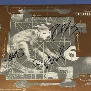 May include: A brown album cover with a monkey illustration in a grid, with the band name "Pixies" and the album title "Doolittle". The cover is signed in black marker. The album art has a vintage aesthetic.