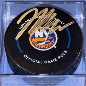May include: Black official game puck with the New York Islanders logo in a clear plastic case. The puck has a gold signature across the top. The logo is orange, blue, and white. The text "NATIONAL HOCKEY LEAGUE" and "OFFICIAL GAME PUCK" are printed on the puck.