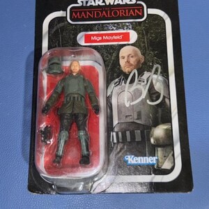 May include: Star Wars: The Mandalorian action figure of Migs Mayfield, packaged on a black and red card. The figure is in a clear plastic case. The packaging features the Star Wars logo and the Kenner brand.