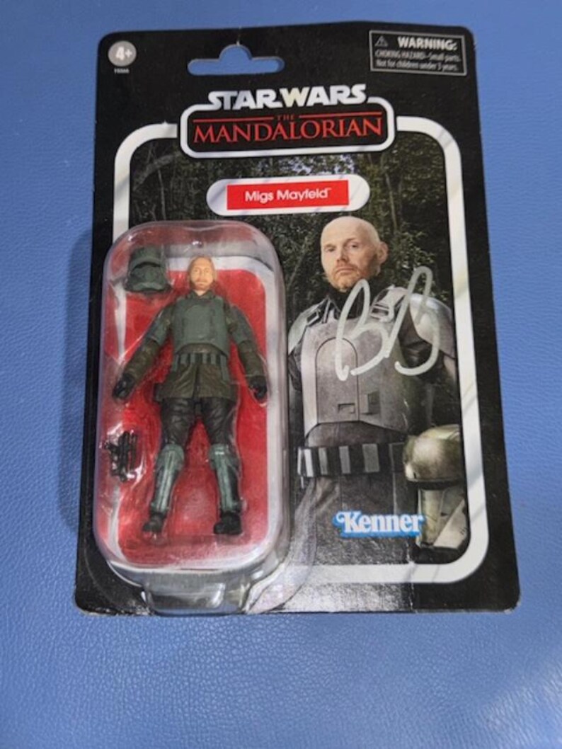 May include: Star Wars: The Mandalorian action figure of Migs Mayfield, packaged on a black and red card. The figure is in a clear plastic case. The packaging features the Star Wars logo and the Kenner brand.