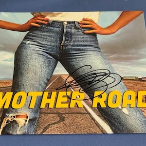 Può includere: Una copertina di album quadrata che mostra una persona che indossa jeans in denim blu con un ginocchio consumato, in piedi su una strada desertica. Il testo MOTHER ROAD è stampato in grassetto giallo in basso, con una firma a inchiostro nero sulla coscia.
