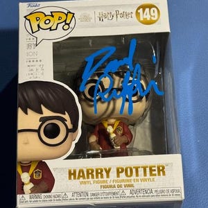 Puede incluir: Una figura de vinilo Funko Pop! de Harry Potter en una caja de exhibición. La figura lleva un uniforme rojo y dorado y gafas. La caja tiene las palabras "Harry Potter" y "149" impresas. La figura está firmada con tinta azul.