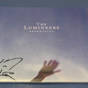 Puede incluir: Portada del álbum "Brightside" de The Lumineers. La portada es un degradado de azul y blanco con el nombre de la banda y el título del álbum en texto blanco. Se ve una sombra de mano. La portada está firmada.