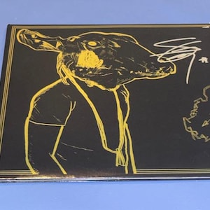 May include: Black square album cover with a yellow illustration of a creature's head and torso. The creature has a long snout and is wearing a shirt. White signature and text are on the right side of the cover.