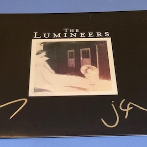 May include: Black album cover featuring the band name "THE LUMINEERS" above a sepia-toned photograph. The photograph shows a person in a white dress and a child. Gold signatures are on the cover.