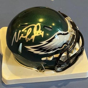 Könnte beinhalten: Ein grüner und silberner Philadelphia Eagles Footballhelm mit schwarzem Gesichtsschutz. Der Helm hat ein weißes und schwarzes Eagles-Logo an der Seite und eine goldene Unterschrift. Der Helm wird auf einem weißen Ständer präsentiert.