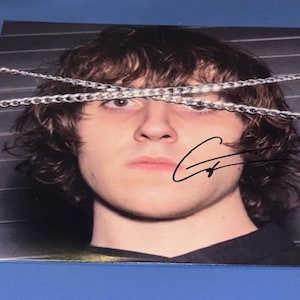 May include: A square print featuring a person with brown hair and a serious expression. Silver chain strands cross the face. The print is signed with a black marker. The background has horizontal gray lines.