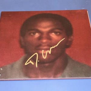 Op de afbeelding: Albumhoes met een portret van een man op een rode achtergrond. Een gouden handtekening is rechtsonder zichtbaar. Het gezicht van de man is enigszins wazig. Een Parental Advisory-label staat linksonder.