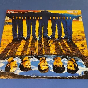 May include: A vinyl record cover for Split Enz titled Conflicting Emotions, featuring a desert landscape with five figures standing in the sun, their shadows cast over a puddle reflecting their faces, with a black ink signature across the center.