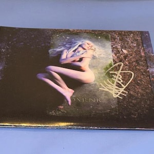 May include: A rectangular print featuring a nude person with long blonde hair, lying on a stone surface. The print has a signature in the upper right corner and the word "OXNRNR" at the bottom.