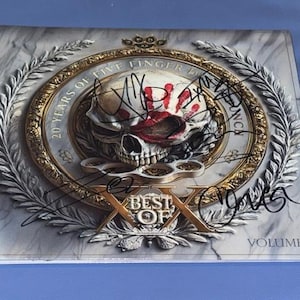 May include: A square music album cover featuring a central skull graphic with a red handprint, brass knuckles, and laurel wreaths. The text reads 20 YEARS OF FIVE FINGER DEATH PUNCH, BEST OF, and VOLUME I. The cover is signed with black ink markers.