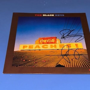 Puede incluir: Una funda cuadrada de disco de vinilo para The Black Keys con una fotografía de un letrero de Coca-Cola sobre un letrero amarillo que dice PEACHES! con dos firmas en tinta negra en la parte frontal. La portada tiene un borde marrón oscuro.