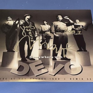 以下が含まれることがあります： モノクロのアルバムカバー。バンドDevoが写っています。バンドメンバーは台座の上に立っています。アルバムタイトルは「Somewhere with Devo」で、「Live at the Palace 1988 + Remix Set」というテキストが含まれています。