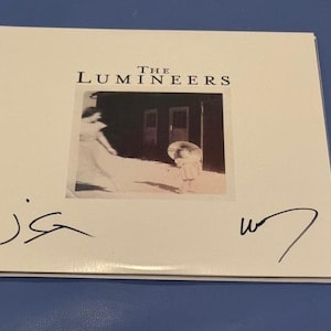 Puede incluir: Caja de CD blanca para el álbum "The Lumineers", con una imagen en sepia de dos figuras. La caja tiene dos firmas y está sobre un fondo azul.
