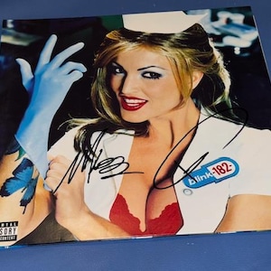 Puede incluir: Portada del álbum con una mujer con uniforme de enfermera, sujetador rojo, guantes azules y un tatuaje de mariposa. La portada del álbum tiene el texto "blink-182" y está firmada. Advertencia Parental Advisory Explicit Content visible.