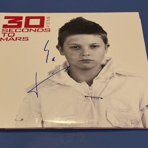 May include: A CD cover for the band 30 Seconds to Mars. The album cover features a black and white photo of a person wearing a white jacket. The band's name is printed in red at the top left. The cover is signed in blue ink.
