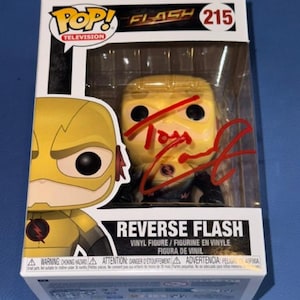 May include: A Funko Pop Television Reverse Flash vinyl figure, number 215, encased in its original window box packaging. The yellow character figure features a red signature across the clear plastic front panel of the box.