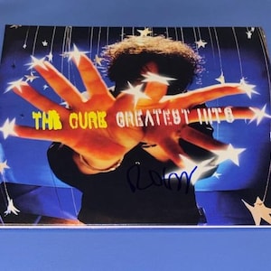 May include: Album cover for "The Cure Greatest Hits" featuring a person's hand with star accents. The text "THE CURE GREATEST HITS" is in yellow. The album cover has a blue background and a signature.